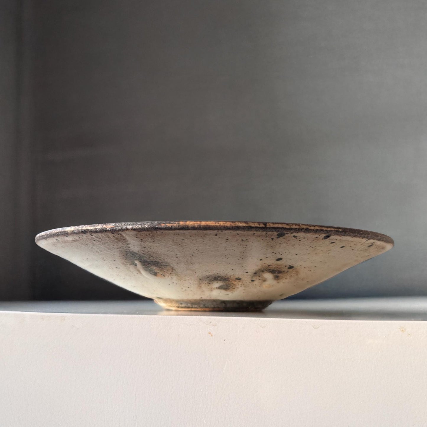 A handcrafted, shallow ceramic bowl with a subtle, earthy tone and a speckled glaze, evoking a sense of understated beauty.