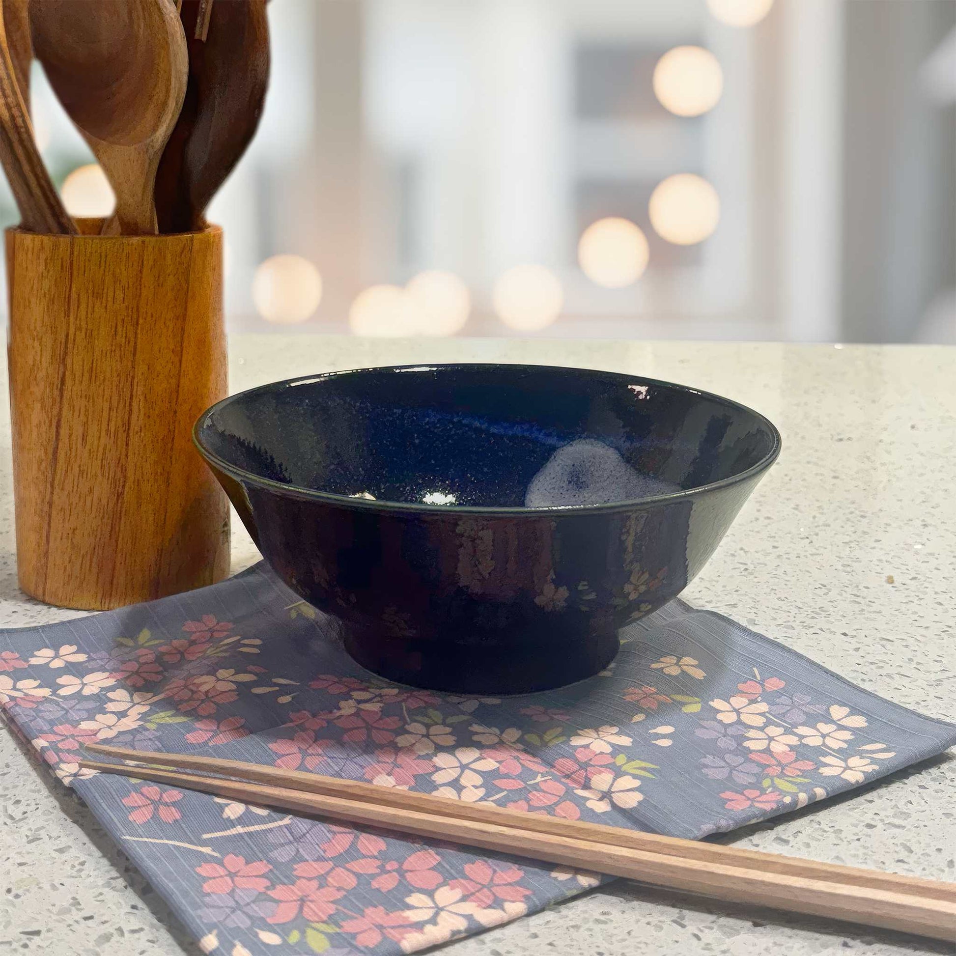 A dark blue, hand-painted Mino ware ramen bowl with a speckled glaze, placed on a floral-patterned placemat.