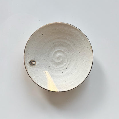 A handcrafted, shallow ceramic bowl with a subtle, off-white glaze and a small, circular indentation in the center.