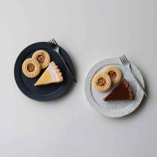 A small, handcrafted plate with a black and white speckled glaze, served with a fork and two types of pie.