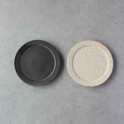 A small, handcrafted plate in black and white, with a speckled glaze, set against a neutral background.