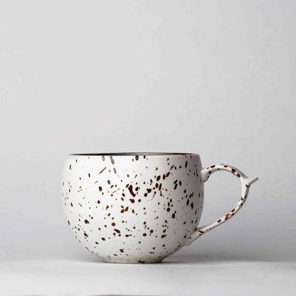 A white, speckled, handcrafted ceramic mug with a handle, set against a plain white background.