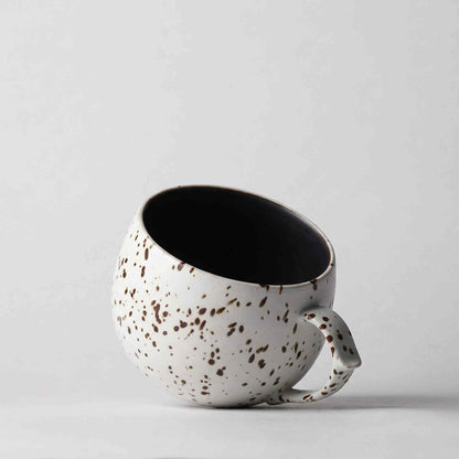 A handcrafted, black and white, speckled ceramic mug with a handle, set against a plain white background.