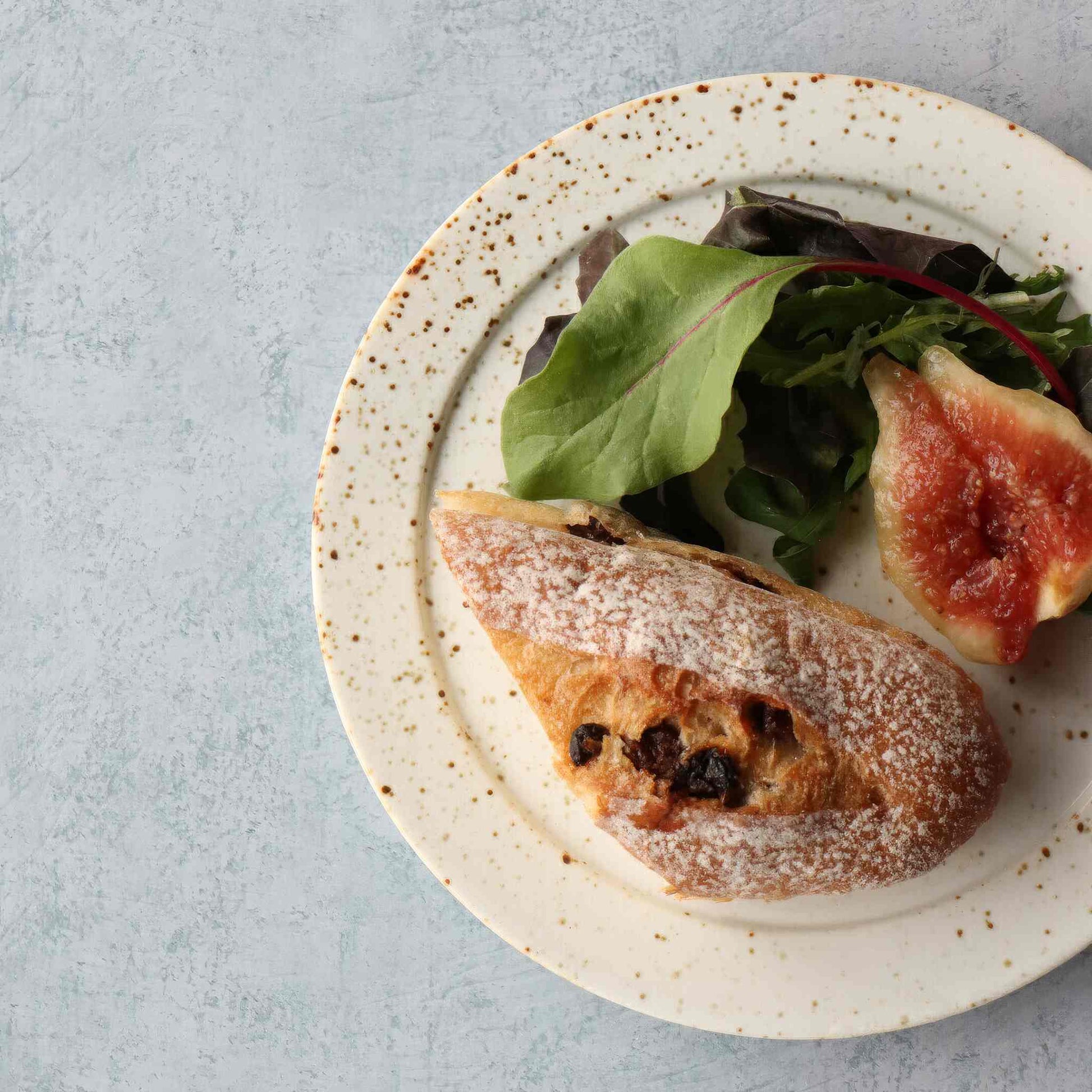 A small, handcrafted plate with a black and white speckled glaze, served with a piece of bread and a salad on a light grey surface.