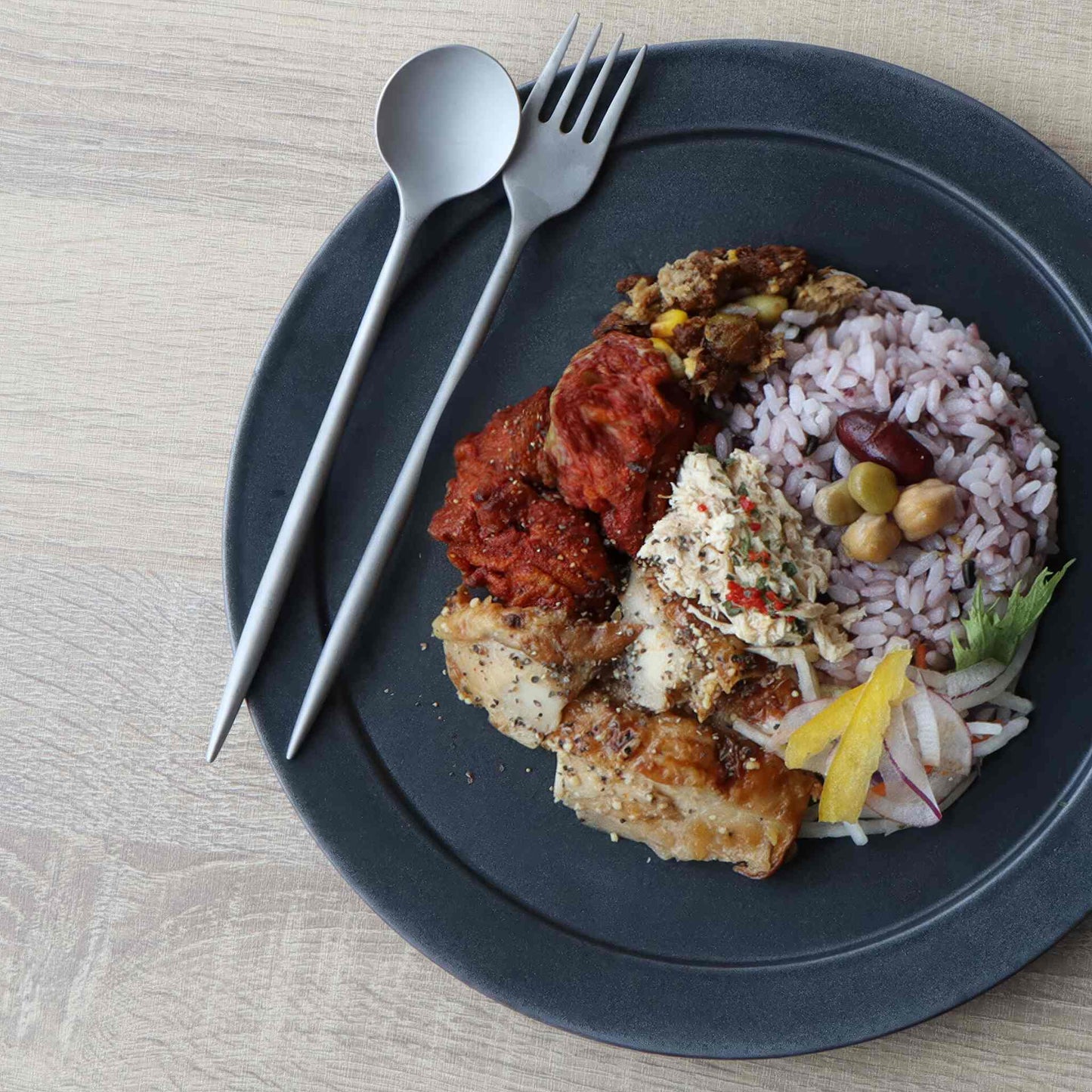 A plate of food, including rice, vegetables, and meat, served on a black plate with a fork and spoon.