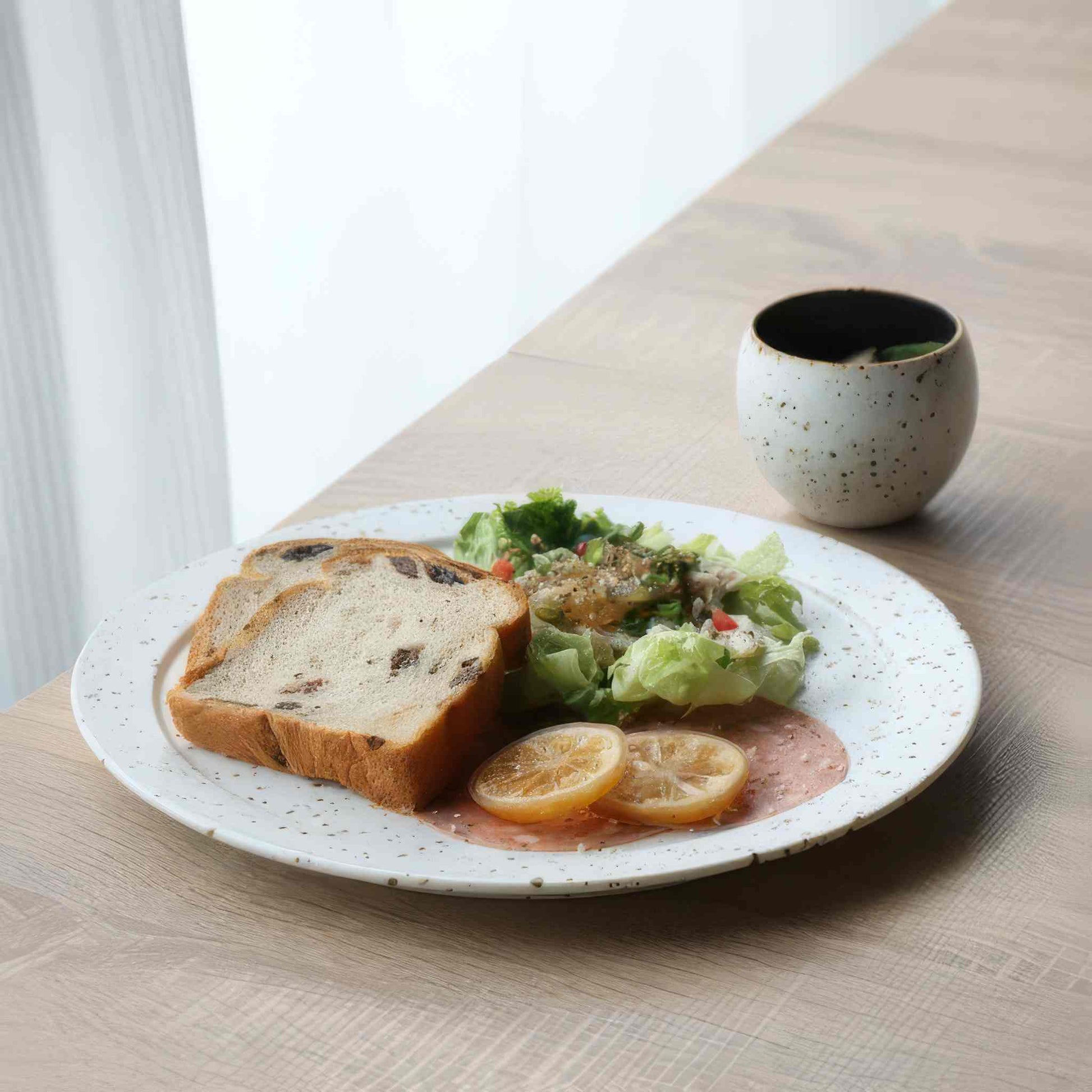 A plate of food, including a slice of bread, a salad, and a slice of lemon, served on a white plate with a speckled pattern.