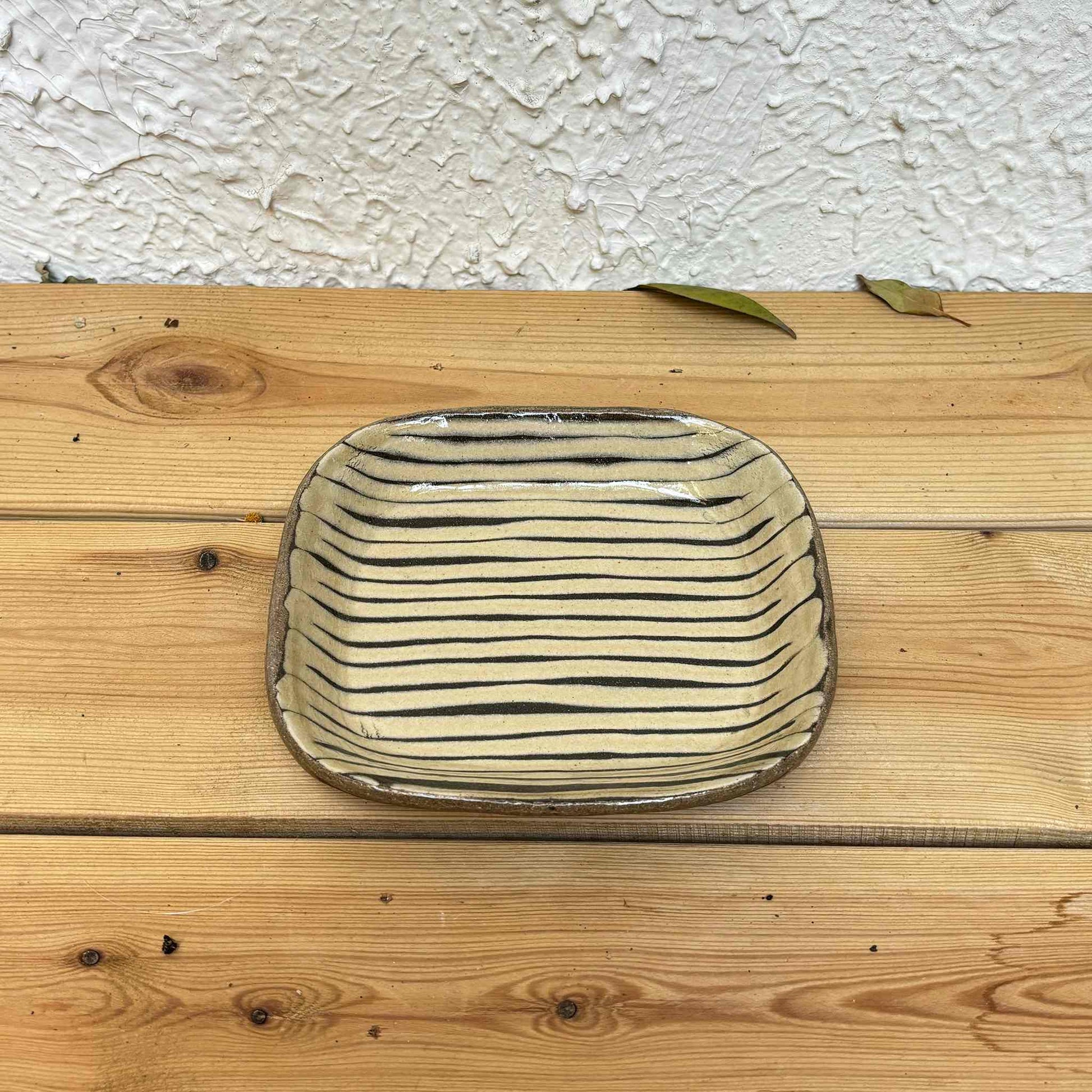 Handmade slipware square plate with unique, hand-applied surface patterns in a neutral, earthy tone, set on a natural wood surface.