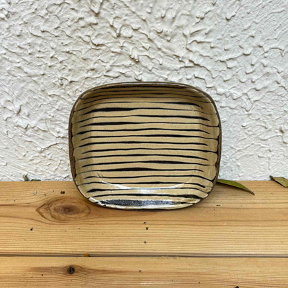 Handmade square plate with hand-applied surface patterns in a neutral color, placed on a wooden surface.