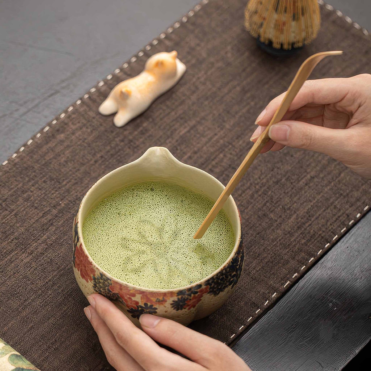 A hand holds a beautiful, hand-painted matcha bowl with a wooden spoon, set on a brown woven mat.