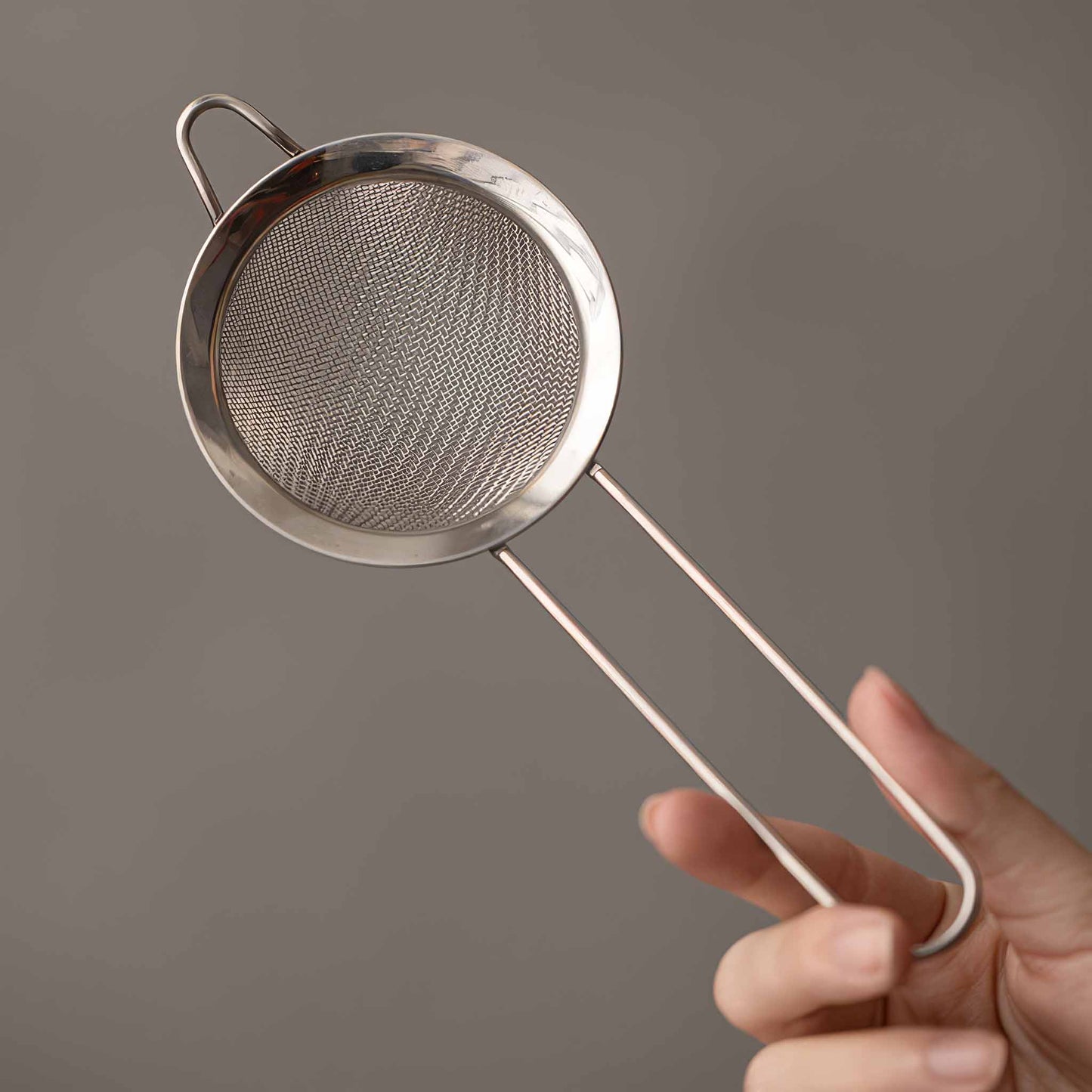 A hand holds a small, delicate, and intricately designed metal strainer with a long handle, against a neutral background.