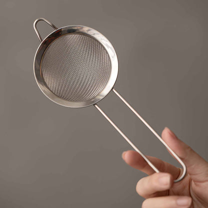 A hand holds a small, delicate, and intricately designed metal strainer with a long handle, against a neutral background.