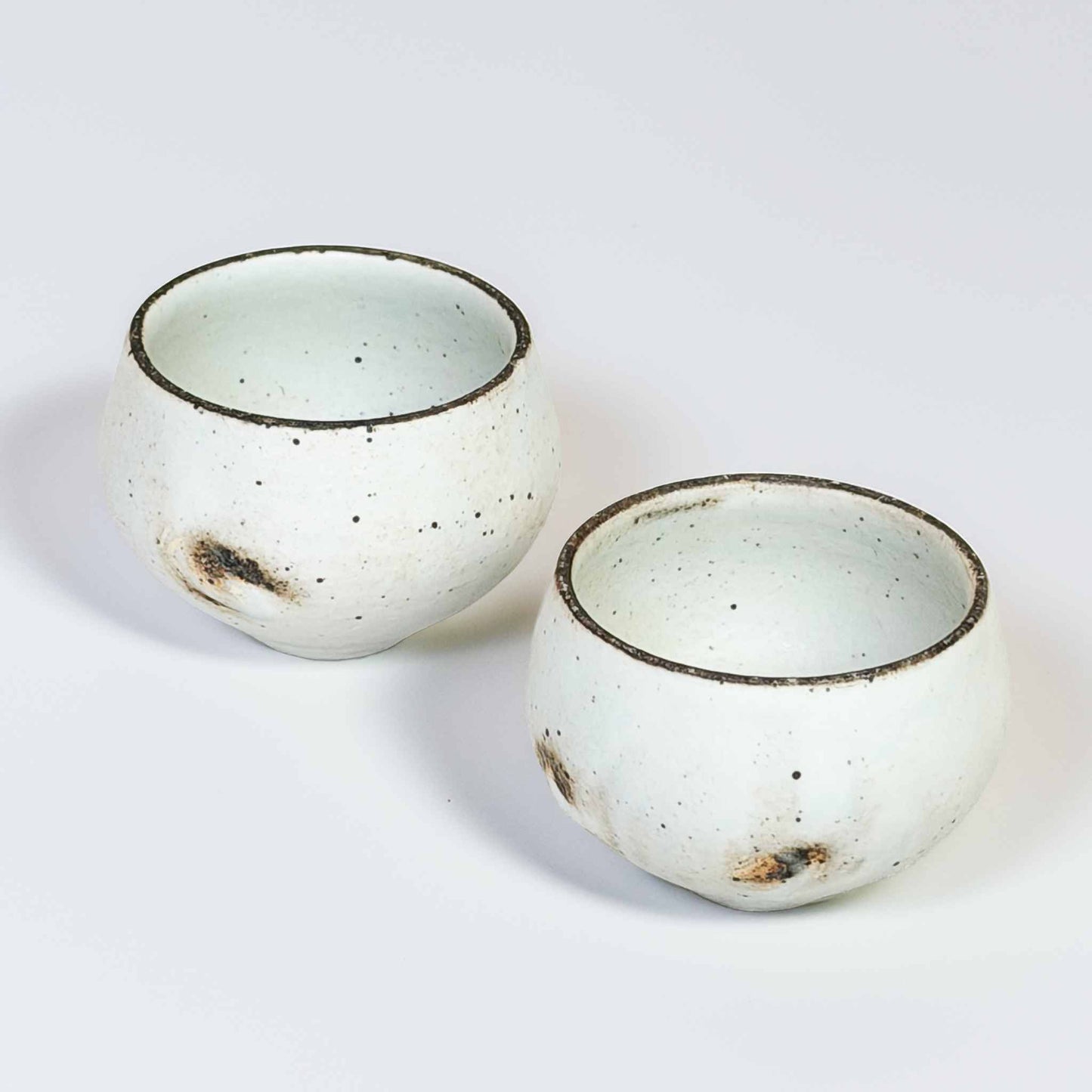 A pair of white, speckled, handcrafted soup cups with a natural glaze, set against a neutral background.