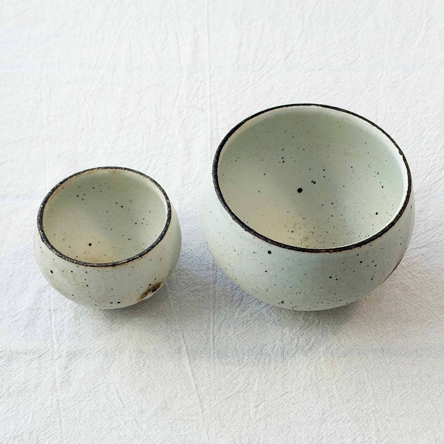 A small, white, speckled ceramic bowl and a larger, white, speckled ceramic bowl sit on a white surface.