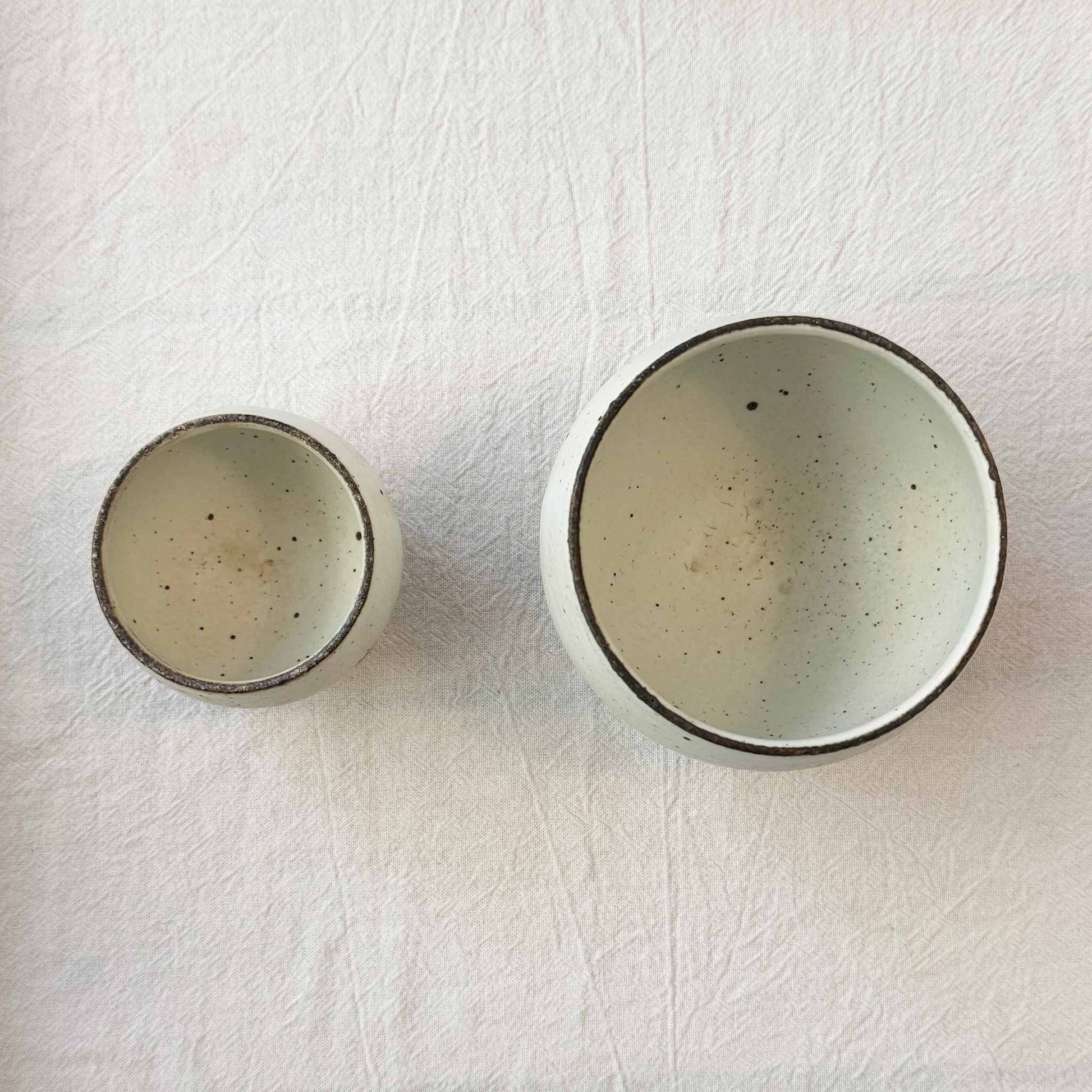 A small, handcrafted white ceramic cup with a speckled glaze, next to a larger version, both set on a neutral background.