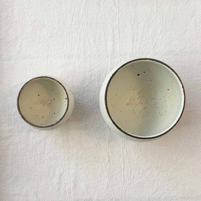 A small, handcrafted white ceramic cup with a speckled glaze, next to a larger version, both set on a neutral background.